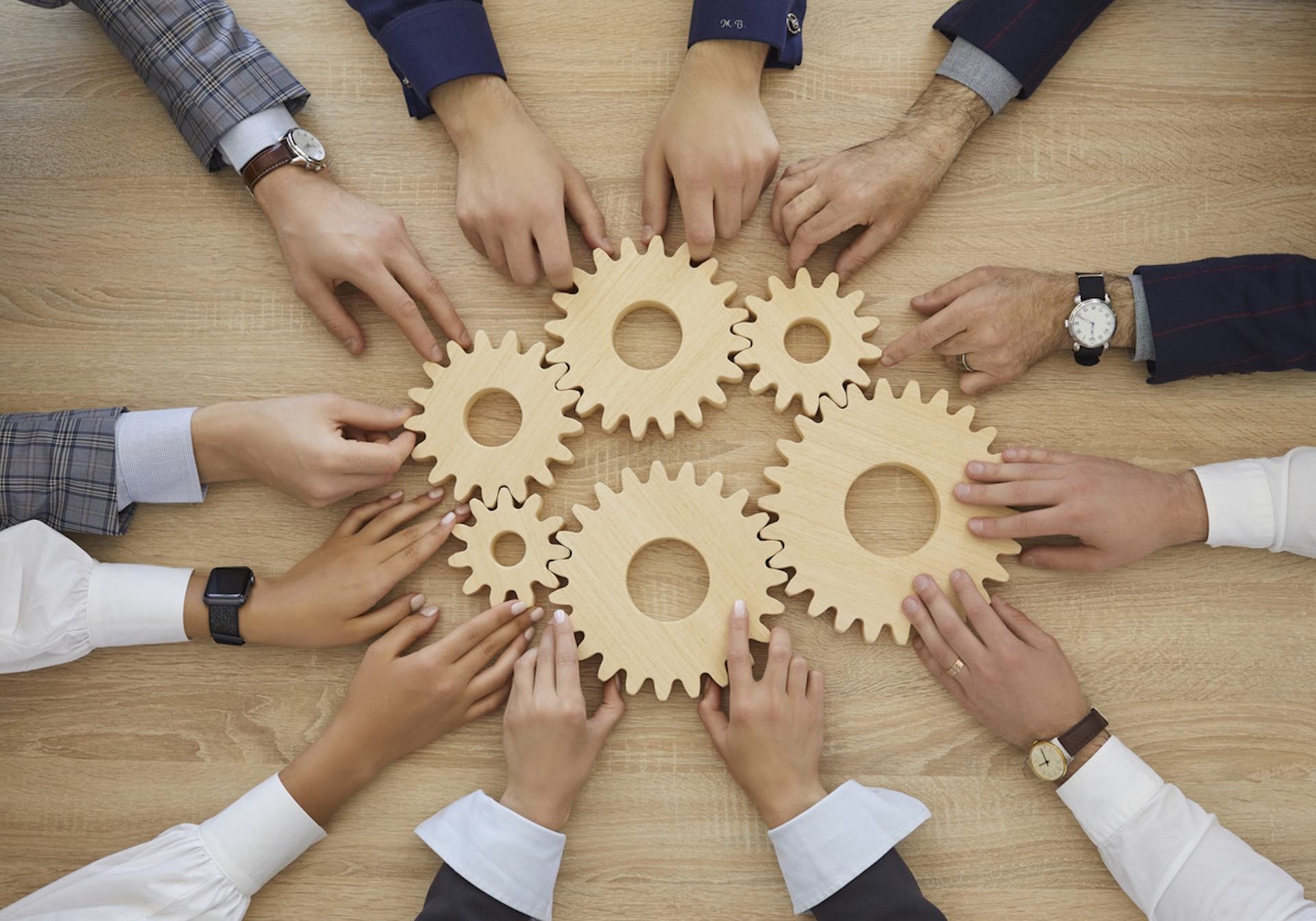 Team of business people join gearwheels. High angle, overhead view of circle of hands holding cogs on office table. Metaphor for good effective business system, cooperation, teamwork and efficiency